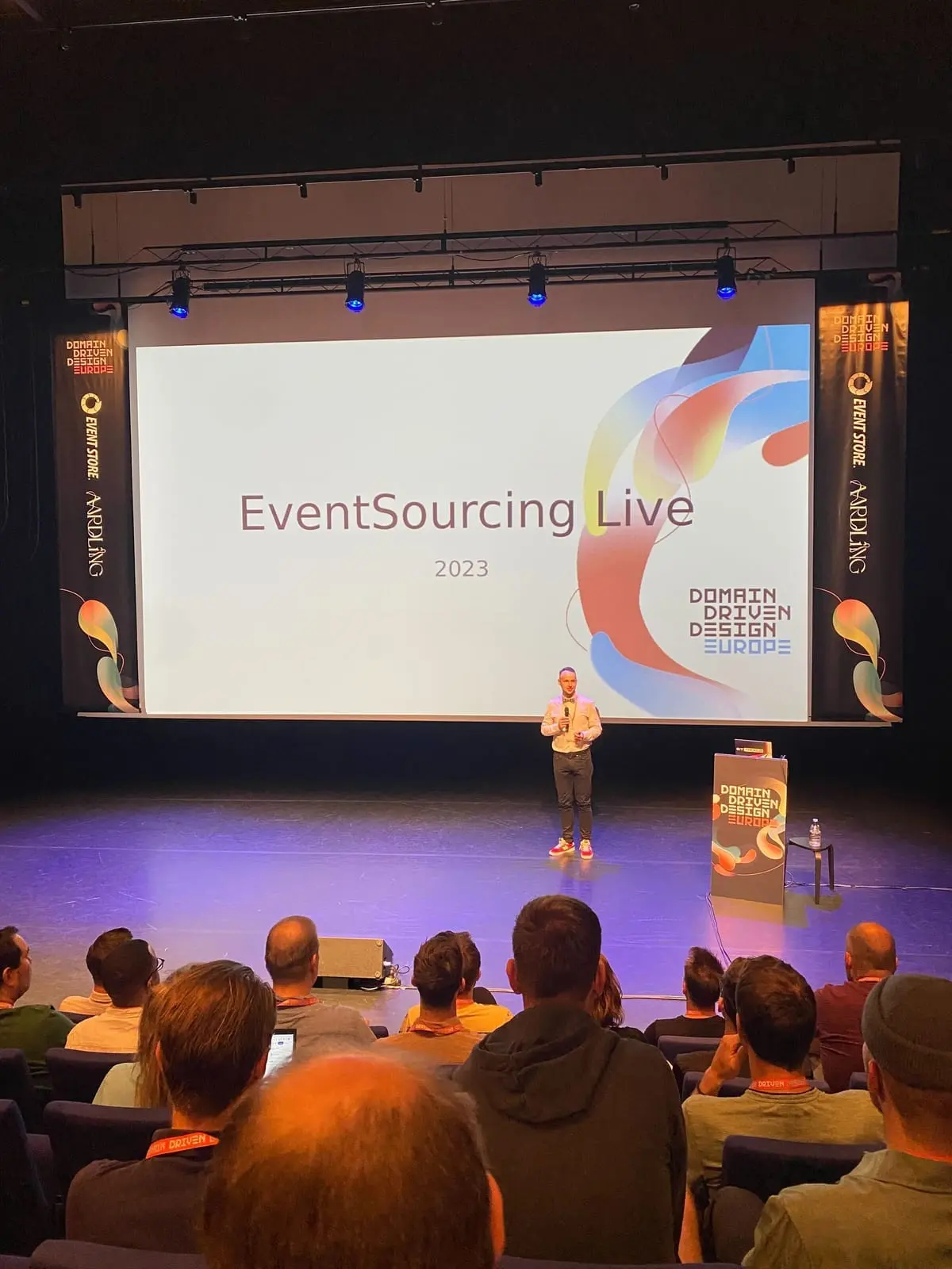 En foredragsholder på scenen foran lysbildet EventSourcing Live 2023, med publikum i salen