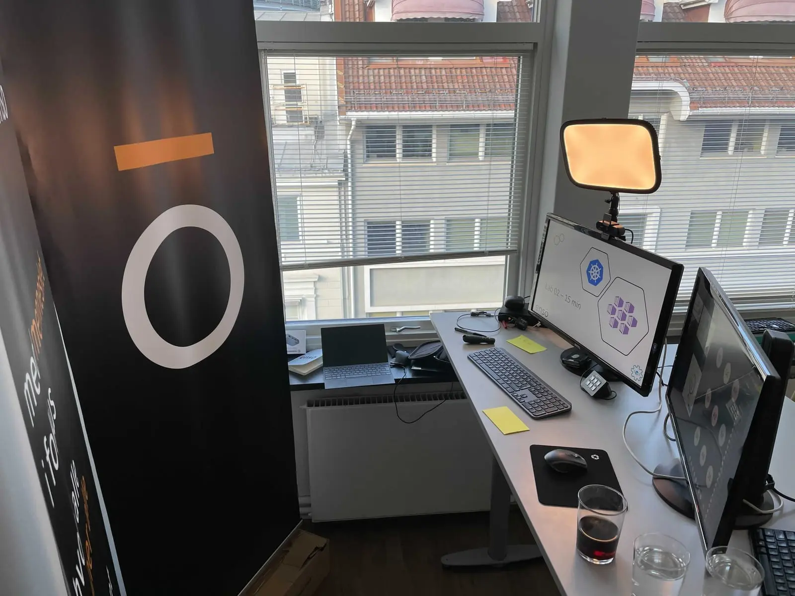 En workshop-arbeidsplass med to skjermer, videolys og en Ensō-roll-up ved vinduet
